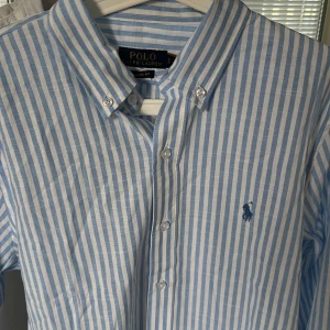 Blåvit randig skjorta från Polo Ralph Lauren - Säljer en klassisk blåvit randig skjorta från Polo Ralph Lauren i storlek S. Skjortan har button down-krage, broderad logga på bröstet och knäppning framtill. Tillverkad i tunn bomull, perfekt för en clean och fräsch stil.