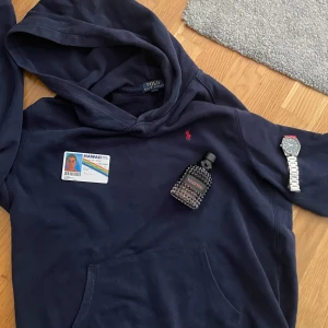 Mörkblå hoodie från Polo Ralph Lauren - Mörkblå hoodie från Polo Ralph Lauren med klassisk röd broderad logga på bröstet. Tröjan har huva och känguruficka framtill. Perfekt för dig som gillar en clean och tidlös stil. Materialet är mjuk bomull som känns skönt mot huden.  Passar, 170/88, (xl barnstorlek). Självklart ordentligt tvättad innan, Vid frågor, tveka inte!😊