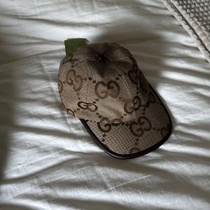 Gucci keps med GG-monogram - Snygg keps från Gucci i beige och brunt med det klassiska GG-monogrammet över hela kepsen. Kepsen har mörkbruna kanter och detaljer, samt justerbar rem bak. Tillverkad i canvas med insida i ljus textil. Perfekt för dig som vill sticka ut med lyxig streetstyle. Helt ny, storlek M!