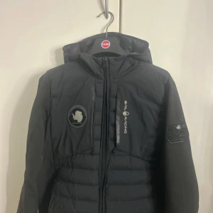 Svart Sail Racing dunjacka med huva - Svart dunjacka från Sail Racing med huva och snygga loggor på bröstet och ärmen. Jackan har quiltad design, dragkedja framtill och en clean look som passar perfekt till kalla dagar. Märket och detaljerna ger jackan en sportig vibe.