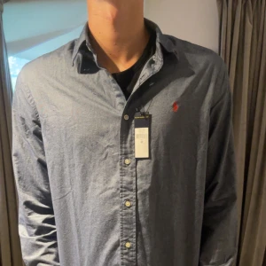 Blå skjorta från Ralph Lauren M - Snygg blå skjorta från Ralph Lauren i klassisk modell med knappar framtill och broderad röd logga på bröstet. Skjortan har lång ärm och är tillverkad i mjuk bomull. Perfekt för dig som gillar stilrena och tidlösa plagg.