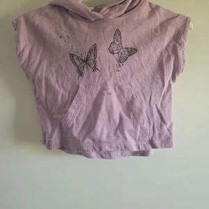 Ljuslila hoodie med fjärilar Åhléns - Kortärmad ljuslila hoodie från Åhléns i 100% ekologisk bomull. Framtill finns ett tryck med två fjärilar och en magficka. Tröjan har huva och en avslappnad, loose passform. Perfekt för dig som gillar söta detaljer och sköna material.