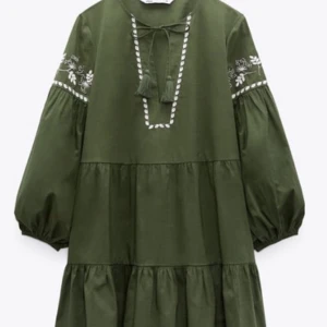 Grön boho-klänning med broderi - Säljer en grön boho-inspirerad klänning med broderade vita detaljer på ärmar och runt halsen. Klänningen har volangkjol, puffiga långärmar och snörning med tofsar vid halsen. Perfekt för dig som gillar avslappnad och trendig stil.