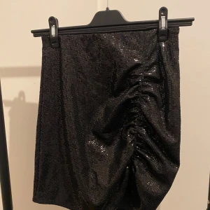 Svart glittrig kortkjol från H&M - Snygg svart kortkjol från H&M med glittriga paljetter och en cool rynkad detalj på ena sidan. Kjolen har en slim passform och är perfekt för att sticka ut med lite extra bling. Materialet är syntet och kjolen är riktigt bekväm att bära.