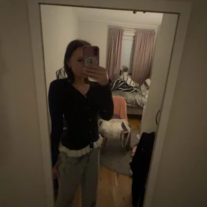 Svart kofta med volang från SHEIN - Svart kofta från SHEIN i storlek S med lång ärm och knappar framtill. Koftan har en snygg volangdetalj i beige vid nederkanten som ger en cool kontrast. Passformen är slim och materialet känns mjukt och stretchigt, perfekt att styla med jeans.