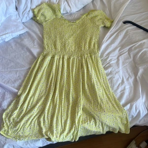 Gul blommig kortklänning Stories M - Supersöt gul kortklänning från Stories med små vita och svarta blommor. Klänningen har smockad överdel, korta ärmar med volang och en flowy kjol. Perfekt för varma dagar och ger en riktigt somrig vibe!