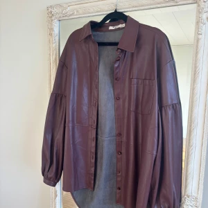 Brun overshirt i skinn från NA-KD - Säljer en oversized brun overshirt i skinnimitation från NA-KD. Skjortan har klassisk krage, knappar framtill, bröstficka och ballongärmar. Perfekt att slänga över en t-shirt för en cool look.