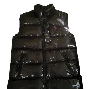 Svart dunväst från Polo Ralph Lauren - Glansig svart puffer dunväst från Polo Ralph Lauren med röd broderad logga på bröstet. Västen har hög krage, två dragkedjefickor och RL-detalj på dragkedjan. Perfekt för lager-på-lager och riktigt snygg till grisch.