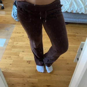 Bruna Juicy Couture  - Mysiga bruna mjukisbyxor från Juicy Couture i velour med snörning i midjan och fickor med lock bak. Byxorna har en relaxed passform och klassisk Juicy-stil, perfekta för chill dagar eller när du vill vara extra comfy men fortfarande snygg. 