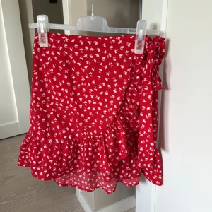 Röd blommig omlottkjol med volang - Superfin röd kort omlottkjol med vita blommor och volangkant. Kjolen knyts i sidan med en söt rosett. Tunn och lätt i materialet, perfekt för sommaren. 