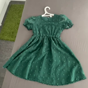 Grön kortklänning med puffärm - Säljer en söt grön klänning med puffiga kortärmar och rund hals. Klänningen har ett diskret blommigt mönster och markerad midja som ger en fin siluett. Perfekt för dig som gillar romantisk och lekfull stil.