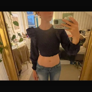 Svart croppad prickig blus med volang - Säljer en svart croppad blus med prickigt mönster och stora volanger på axlarna. Toppen har långärmade puffärmar och rund halsringning. Materialet känns lätt och har struktur, perfekt för dig som gillar statement-plagg.