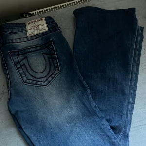 True Religion blå jeans med röd söm - Säljer ett par snygga blå jeans från True Religion med ikonisk röd kontrastsöm och stor broderad logga på bakfickan. Klassisk femficksmodell med silverfärgade knappar och läderpatch bak. Jeansen har en rak passform och är tillverkade i slitstarkt denim. Säljer då de är lite för stora för mig. Jeansen är uppsydda och passar i längden på mig som är 165. De är i storlek 27