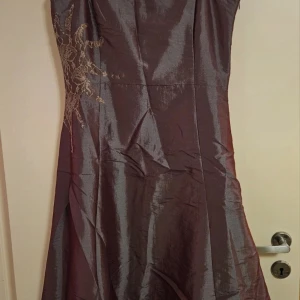 Lila festklänning från Vila - Snygg lila festklänning från Vila med axelbandslös design och spetsdetaljer på sidan. Klänningen har en glansig satinliknande yta och en elegant, figurnära passform upptill som faller ut i en vidare kjol. Perfekt för bal eller fest!