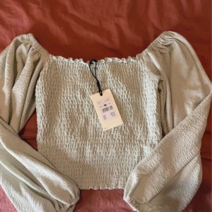 Ljusgrön blus med puffärmar - Supersöt ljusgrön blus med smockad kropp och vida puffärmar. Offshoulder-modell med elastisk kant upptill och nertill. Perfekt för dig som gillar romantisk och trendig stil.