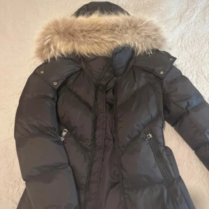 Svart Moncler dunjacka med päls - Snygg svart dunjacka från Moncler med fluffig äkta päls runt huvan och markerat bälte i midjan. Jackan har dragkedjor, två fickor framtill och quiltad design. Perfekt för kalla vinterdagar när du vill vara både varm och stilren.