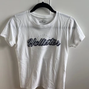 Vit och Blå Hollister t-shirt - Vit t-shirt från Hollister med klassisk rund hals och korta ärmar. Framsidan har ett stort Hollister-tryck i blått med broderad effekt. T-shirten är gjord i mjuk bomull och har en normal passform, perfekt för en chill och stilren look. Använd ett par gånger men inga hål💕💕 Jag tror storleken är M/S