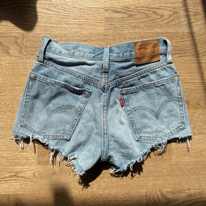 Levi's 501 ljusblå jeansshorts - Levi's 501 jeansshorts i ljusblå denim med råa, fransiga kanter och slitna detaljer. Shortsen har fickor, knappgylf och den ikoniska läderpatchen bak. Shortsen är i storlek w24. 