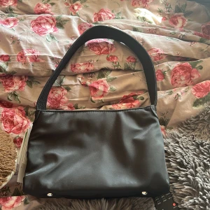 Svart axelväska  - En stilren svart axelväska med bredt, vadderat axelband och enkel design. Väskan har dragkedja upptill och flera fack inuti för smidig organisering. Perfekt storlek för att bära med sig det viktigaste och passar till många olika outfits. Ord pris  995:- 