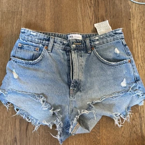 Blå jeansshorts med fransar från Zara - Snygga blå jeansshorts från Zara i klassisk femficksmodell. De har hög midja, slitna detaljer och råa fransade kanter för en cool vibe. Perfekta för sommaren och festivaler. Materialet är robust denim och shortsen har knapp och dragkedja framtill.