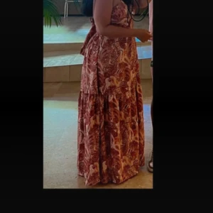 Paisleymönstrad långklänning med knyt - Säljer en bohemisk långklänning med paisleymönster i mörkrosa och ljusrosa toner. Klänningen har omlottkänsla upptill, knyt i nacken och volangdetalj nedtill. Perfekt för sommarens alla tillfällen när du vill sticka ut med färg och mönster. Den har en knyt vid ryggen som går att justera hur du vill att brösten ska sitta. Den är så fin och skön på. Använd enbart engång, bild nummer 1