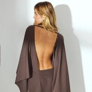 Brun långklänning med öppen rygg - Snygg brun långklänning med dramatisk öppen rygg och vida, svepande ärmar. Klänningen har en elegant och minimalistisk design som ger en cool och sofistikerad vibe. Perfekt för dig som vill sticka ut med en unik siluett och djup rygg.