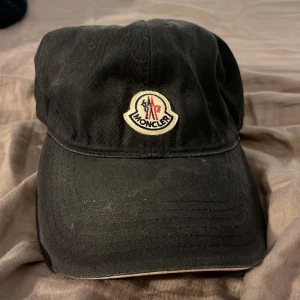 Svart keps från Moncler - Svart keps från Moncler med klassisk böjd skärm och broderad logotyp framtill. Kepsen har en enkel och stilren design, tillverkad i bomull med röd insida och märkning på insidan. Perfekt för dig som gillar exklusiva accessoarer.