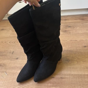 Svarta mockastövlar med klack - Snygga svarta stövlar i mocka med spetsig tå och låg klack. Stövlarna har en slouchy passform och en liten guldfärgad knappdetalj upptill. Det är Pernilla Wahlgrens märke