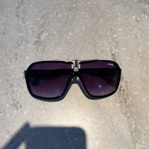 Svarta Carrera solglasögon shield-modell - Snygga svarta Carrera solglasögon med stor shield-formad lins och mörka tonade glas. Ramen är i blank svart plast med silverfärgad detalj vid bryggan och Carrera-logga på skalmarna. Perfekt för dig som vill sticka ut med en sportig och retro vibe.