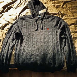 Marin Blå stickad hoodie från Polo Ralph Lauren - Marin blå stickad hoodie från Polo Ralph Lauren med kabelstickat mönster och röd logobrodyr på bröstet. Tröjan har huva med snörning och långa ärmar, perfekt för dig som gillar att se klassisk och stilren ut.