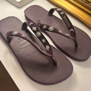 Lila Havaianas sandaler flipflops stjärndetaljer ”top cosmic” - Snygga lila flip-flops från Havaianas med sammetsliknande remmar dekorerade med silvriga stjärnor, månar och planeter. Perfekta för sommaren och strandhäng, med mjuk och bekväm sula i syntetmaterial. Slutsålda överallt, äldre modell som ej går att få tag i. Storlek 37/38.  Nypris: ca 900kr 💜