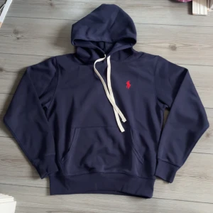 Marinblå hoodie från Polo Ralph Lauren - Snygg marinblå hoodie från Polo Ralph Lauren med klassisk röd logga broderad på bröstet. Tröjan har huva med vit snörning, känguruficka och långa ärmar. Perfekt för en chill och stilren look.