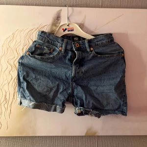 Blå jeansshorts från 157, XS - Klassiska blå jeansshorts från 157 i storlek XS. Modellen har hög midja, fem fickor och råa, upprullade benslut för en avslappnad vibe. Perfekta till sommaren och lätta att matcha med allt. Snygga detaljer med synliga sömmar och knappstängning framtill.