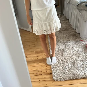 Vit kortkjol med broderad volang - Supersöt vit kortkjol med broderad volangkant och hålmönster. Kjolen har en luftig passform och är perfekt för varma dagar. Den är gjord i mjukt bomullstyg och har en feminin, fräsch vibe som passar till sneakers eller sandaler.