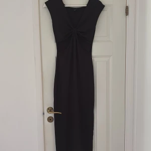 Svart långklänning med knyt - Säljer en svart långklänning  med snygg knytning framtill och cut-out detaljer under bysten. Klänningen har breda axelpartier och är ärmlös, vilket ger en cool och modern siluett. Perfekt för dig som gillar stilrena och trendiga plagg.