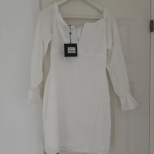 Vit kort klänning med volangärm - Säljer en vit kort klänning med långa ärmar och volangdetalj vid ärmslut. Klänningen har en lätt rynkad kant vid halsen och en liten öppning framtill. Perfekt för dig som gillar en clean och fräsch look med romantiska detaljer.