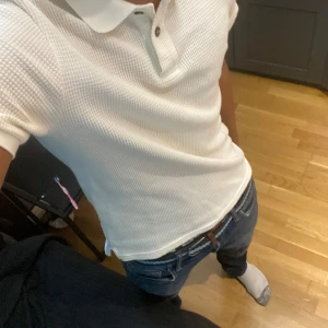 Vit piké t-shirt från H&M - Säljer en stilren vit piké t-shirt från H&M med korta ärmar och klassisk krage. Tröjan har en snygg strukturerad, rutmönstrad yta och knappar vid halsen. Perfekt för en clean och fräsch look.
