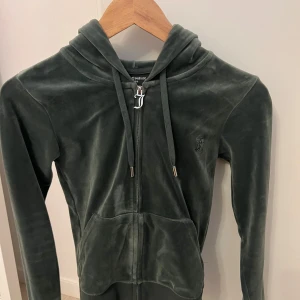 Juicy Couture mörkgrön velour hoodie/byxor - Juicy Couture hoodie samt byxorna i mörkgrön velour med dragkedja och huva. Tröjan har långa ärmar, snörning vid halsen och två fickor framtill. Ikonisk JC-logga broderad på bröstet. Perfekt för chill dagar och en skön, mjuk känsla. Byxorna har kortats ner, jag är 1,52 cm lång. Säljer både koftan och byxorna för 800 kr.