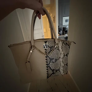 Beige handväska Victoria's Secret - Snygg beige handväska från Victoria's Secret med coolt ormmönster på ena sidan. Väskan har guldfärgade detaljer, kedja och dragkedja upptill. Perfekt storlek för allt du behöver och riktigt trendig look.