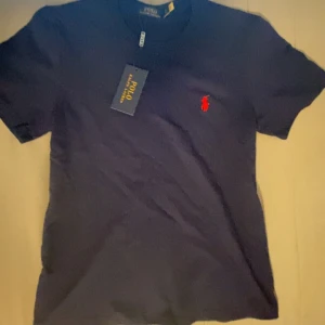 Marinblå t-shirt från Polo Ralph Lauren - Snygg marinblå t-shirt från Polo Ralph Lauren med klassisk röd logga broderad på bröstet. T-shirten har rund hals och korta ärmar, perfekt för en clean och stilren look. Materialet är mjuk bomull som känns skönt mot huden.