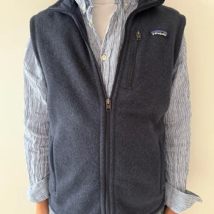 Mörkblå fleeceväst från Patagonia - Snygg mörkblå fleeceväst från Patagonia med hel dragkedja framtill och hög krage. Västen har två sidofickor med dragkedja och en bröstficka med dragkedja. Perfekt att slänga över en skjorta för en chill och sportig look. Nypris 1400kr, hör av er vid frågor!