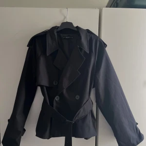 Svart trenchcoat med bälte XS-M - Stilren svart trenchcoat från Bershka med klassisk dubbelknäppning, bred krage och bälte i midjan. Jackan har långa ärmar med knäppdetaljer och axelklaffar för en cool look. Perfekt för dig som vill ha en tidlös och trendig jacka. 