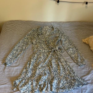 Blommig klänning från ONLY - Säljer en blå och vit omlottklänning från ONLY i storlek 34. Klänningen har lång ärm, volangkant nedtill och knyt i midjan. Använde en enstaka gång. 