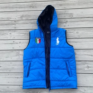 Blå dunväst Polo Ralph Lauren Italia - Snygg blå dunväst från Polo Ralph Lauren med huva och dragkedja. Broderad stor vit logga framtill, italiensk flagga och texten 'Italia' både fram och bak. Quiltad design och två fickor med dragkedja. Perfekt för dig som gillar sportig och cool stil.