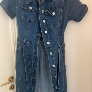 Jeansklänning med bälte och knappar - Snygg jeansklänning i blå denim med korta ärmar, markerad midja och bälte. Klänningen har knappar framtill, bröstfickor med lock och en slits framtill. Figurnära passform som framhäver siluetten och coola sömdetaljer. Helt ny, oanvänt.