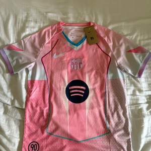 Rosa Barça Ställ (#10 Lamine Yamal) - Snygg FC Barcelona fotbollströja från Nike i rosa med vita och ljusblå detaljer. Kortärmad med V-ringad krage och tryckta ränder. Spotify-logga på bröstet och Lamine Yamal 10 på ryggen. Tillverkad i lätt, ventilerande polyester. Perfekt för dig som älskar Barça. Det har blivit något fel i produktionen så det står att det är en M på tröjan men det är storlek S stor det även på lappen som sitter på tröjan 