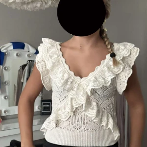 Vit stickad topp med volang och spets - Superfin vit stickad topp med breda volangdetaljer och spets längs v-ringningen och axlarna. Toppen är ärmlös och har ett vackert hålmönster i hela tyget. Perfekt för dig som gillar romantisk och söt stil. Den säljs inte längre 