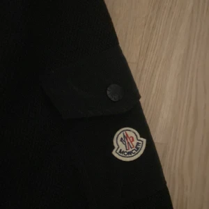 Svart pufferjacka från Moncler M - Snygg svart pufferjacka från Moncler i storlek M. Jackan har quiltad framsida, stickade ärmar och axelficka med Moncler-logga. Dragkedja framtill och diskreta knappar på fickorna. Perfekt för dig som vill ha en stilren och exklusiv look. Pris kan diskuteras och varor kan oxå bytas 
