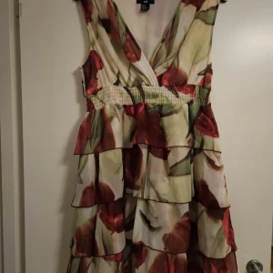 Blommig volangklänning från H&M - Säljer en superfin blommig klänning från H&M i storlek 40. Klänningen har flera lager med volanger, omlott-vringning och ett smockat midjeband. Färgerna går i grön och röd med ett blommigt mönster. Ärmlös och perfekt för sommarens alla tillfällen.