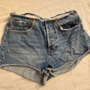 Blå jeansshorts med rå kant - Säljer ett par blå jeansshorts med hög midja och rå, fransig kant upptill och nedtill. Klassisk femficksmodell med slitna detaljer och grova sömmar. Perfekt för dig som gillar en edgy och avslappnad stil under sommaren. STOR I STORLEKEN